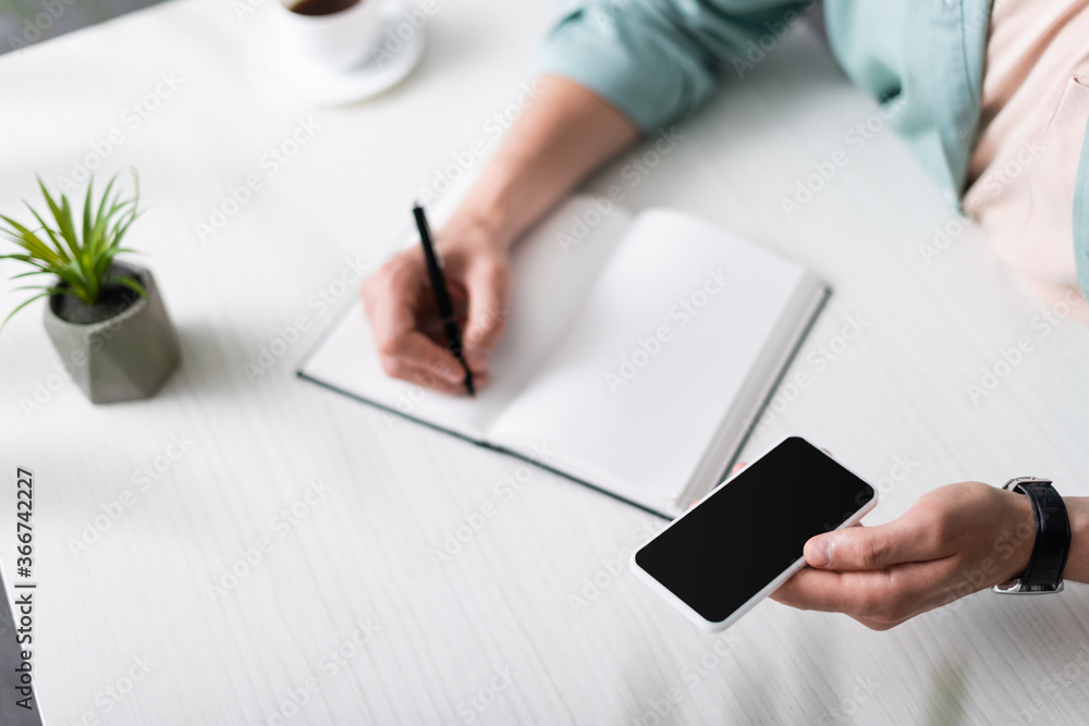 Cropped view of man holding smartphone with blank screen while writing on notebook at table, earning online concept Cropped view of man holding smartphone with blank screen while writing on notebook at table, earning online concept