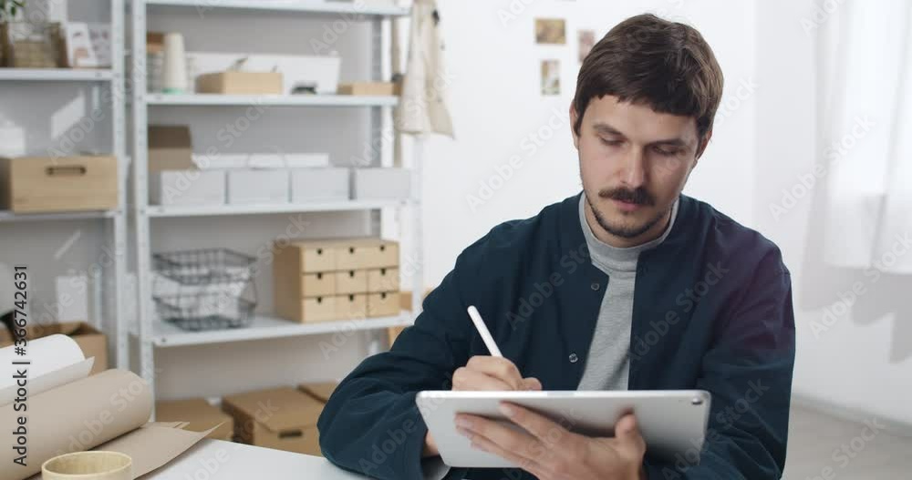 Young man holding notepad and drawing with stylus on it. Creative guy with moustache holding digital tablet while creating illustration and sitting at cosy workplace.Concept of art Young man holding notepad and drawing with stylus on it. Creative guy with moustache holding digital tablet while creating illustration and sitting at cosy workplace.Concept of art