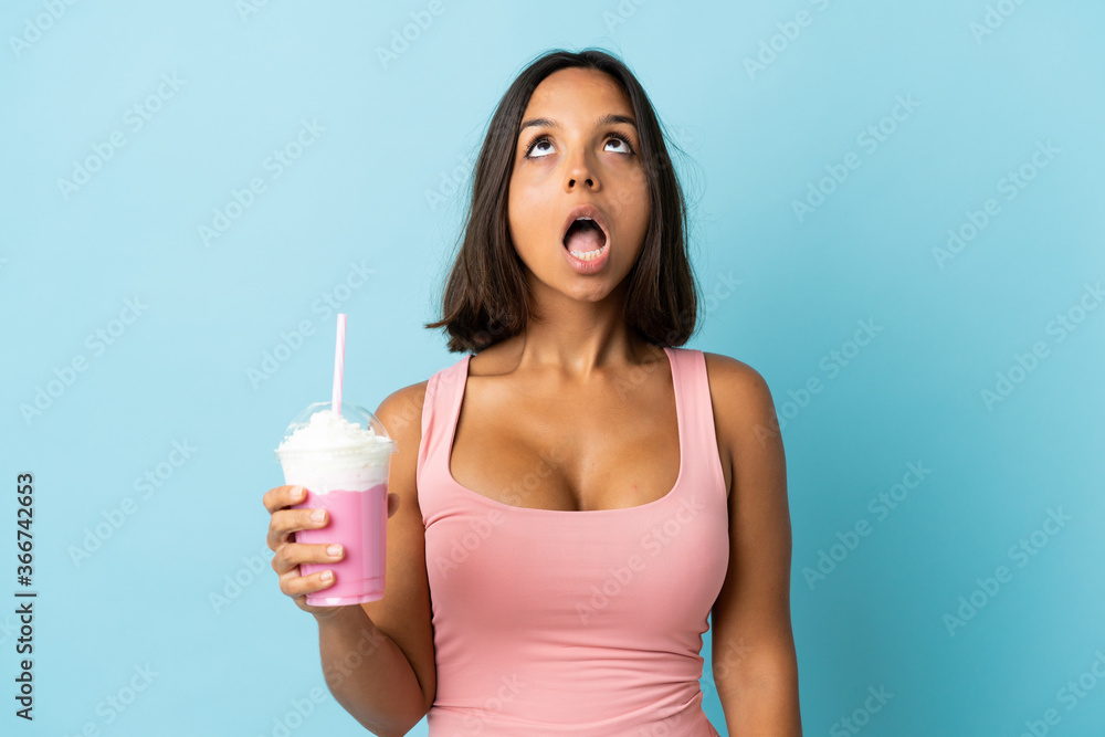 Young woman with strawberry milkshake isolated on blue background looking up and with surprised expression Young woman with strawberry milkshake isolated on blue background looking up and with surprised expression