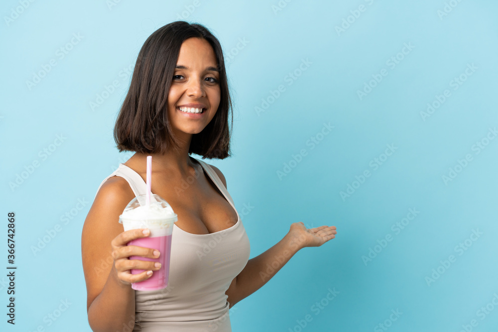 Young woman with strawberry milkshake isolated on blue background extending hands to the side for inviting to come Young woman with strawberry milkshake isolated on blue background extending hands to the side for inviting to come