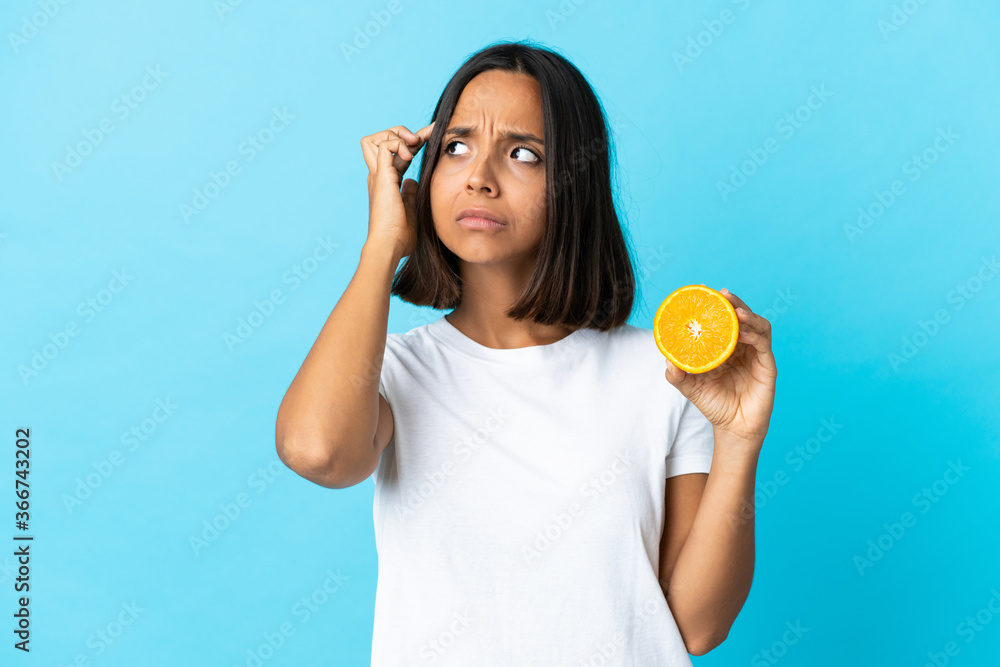 Young asian girl holding an orange isolated on blue background having doubts and thinking Young asian girl holding an orange isolated on blue background having doubts and thinking