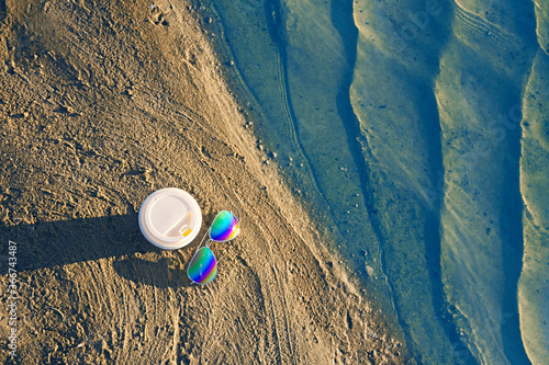 Sunglasses with colored glasses and disposable cup on sand, sea beach and travel concept