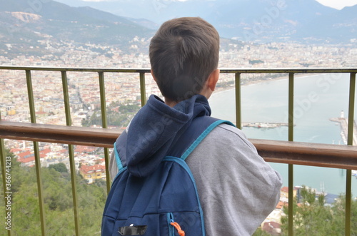 Little boy, looking at the amazing view on the Mediterranian sea gulf with touristic ships and aincient castle and a city from the mountain with a backpack on the back photo