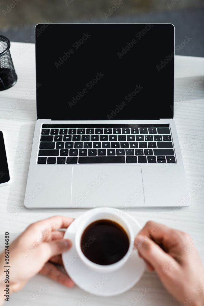 Cropped view of man holding coffee cup near laptop with blank screen, earning online concept Cropped view of man holding coffee cup near laptop with blank screen, earning online concept
