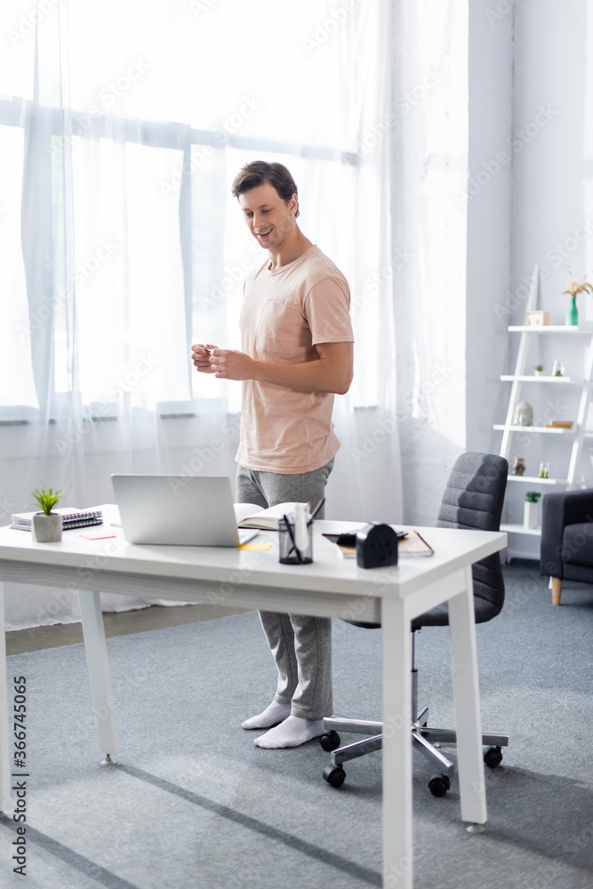 Smiling freelancer standing in front of laptop on table with stationery at home, earning online concept
