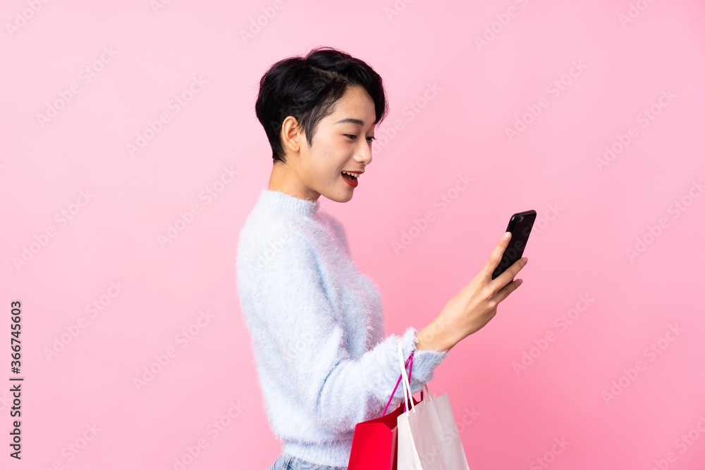 Young Asian girl over isolated pink background holding shopping bags and writing a message with her cell phone to a friend Young Asian girl over isolated pink background holding shopping bags and writing a message with her cell phone to a friend