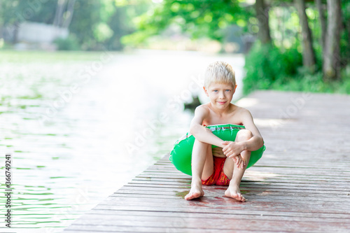 smiling blond boy sits on a pier near the lake with a circle, swims, a child learns to swim, a child's rest in the village