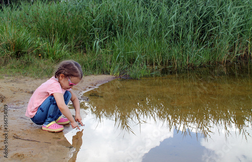 A little girl plays by a pond against the background of green thickets. The concept of a happy childhood and outdoor recreation