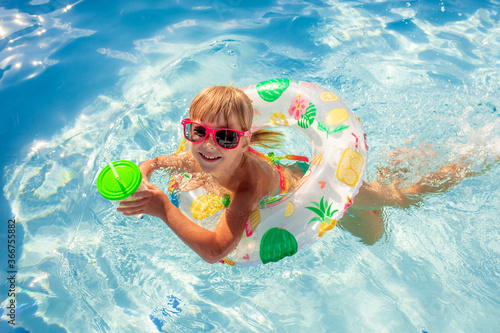 A little girl swims in an inflatable circle in the pool, with a cocktail in her hands