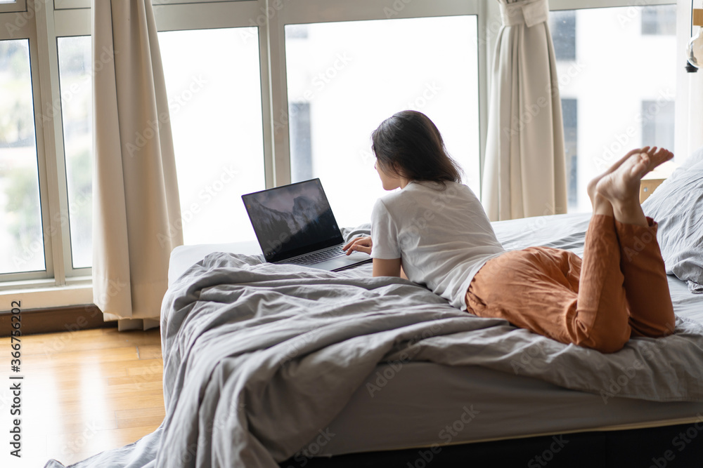 A beautiful young brunette girl is working on the laptop while lying on a bed by a panoramic window. Stylish modern interior. A cozy workplace. Shopping on the Internet.