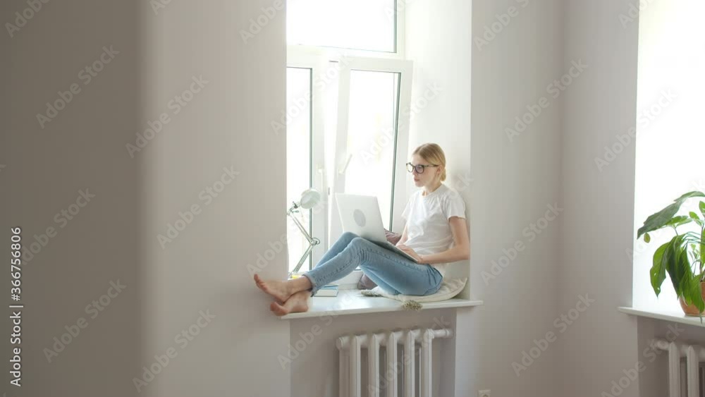 Young woman blogger sitting in room on windowsill in day light calling video call, speaking with friend. Distance learning Young woman blogger sitting in room on windowsill in day light calling video call, speaking with friend. Distance learning