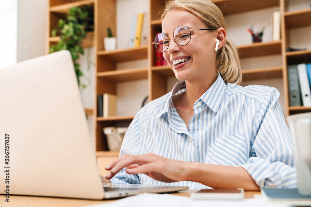 Image of woman using earphones and laughing while working with laptop Image of woman using earphones and laughing while working with laptop