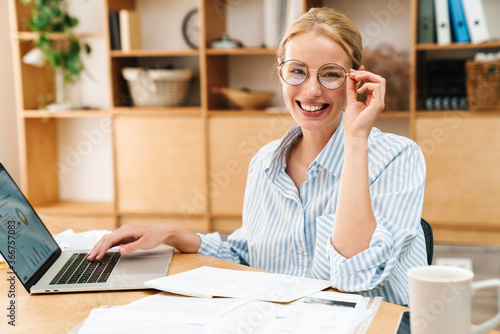 Image of cheerful woman in eyeglasses working with papers and laptop