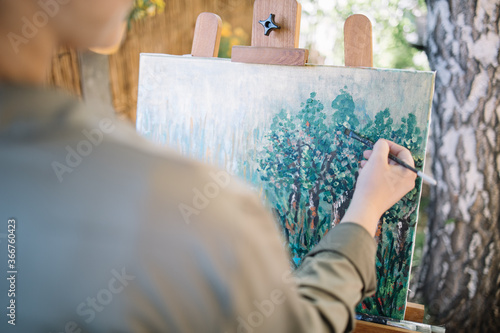 Hand with paintbrush painting picture in nature. Female hand holding paintbrush and painting on canvas outdoor.