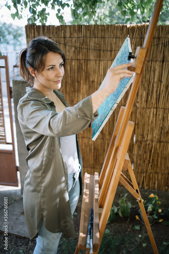 Beautiful woman taking painting from a tripod outdoor. Portrait of pretty girl in theyard looking at canvas stand while getting painting from it.