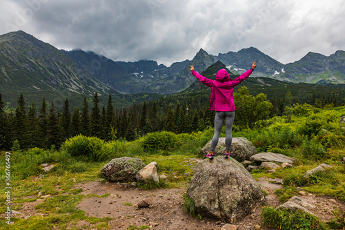 Hooded woman looking at the Tatra mountain panorama in the rain with hands in victory pose - horizontal orientation