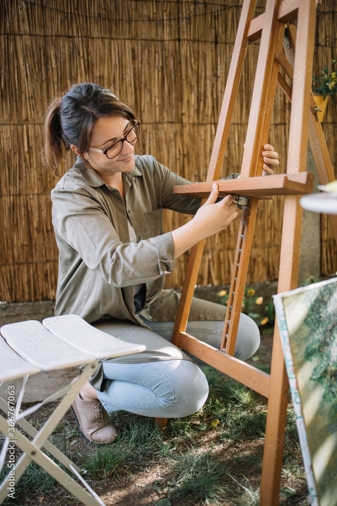 Smiling woman preparing tripod for placing canvas. Female artist preparing wooden tripod for adding painting in garden workshop.