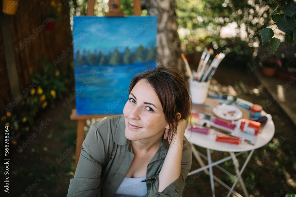 High angle view of woman artist sitting in outdoor art studio. Portrait of woman painter posing in garden art studio with tripod, canvas, picture and paint brushes.