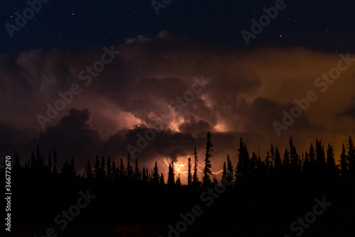 Lightning bolt at night in storm clouds on mountain with trees below starry sky