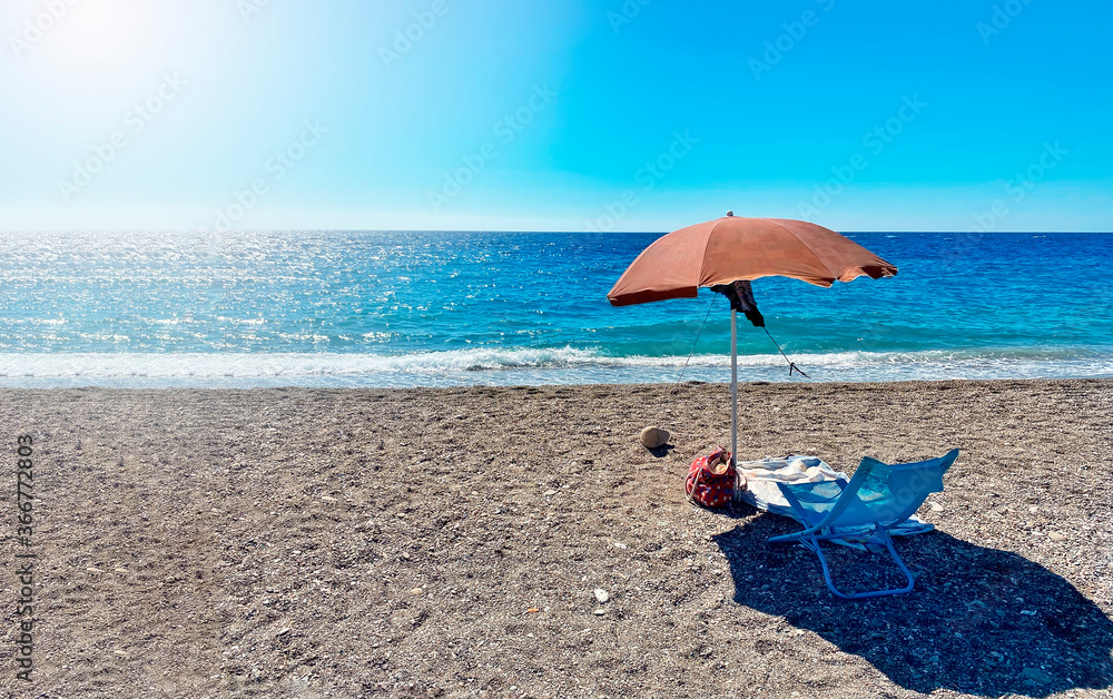 A deckchair under an umbrella facing the blue sea in a stony beach in Sicily, Italy