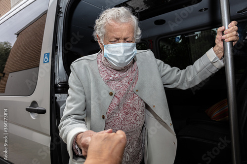 An elderly woman wearing a medical mask gets out of a disabled car.
the concept of transporting patients in the car.