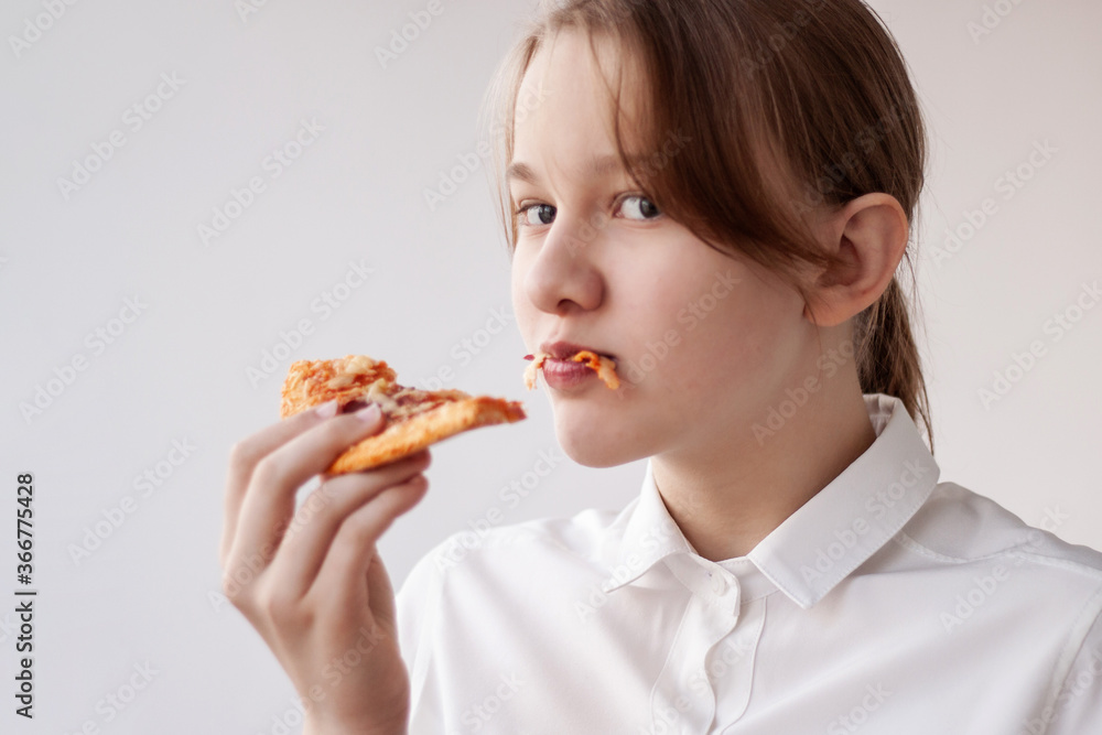 A teenage girl in a white shirt has taken a bite of pizza and is holding it in her mouth