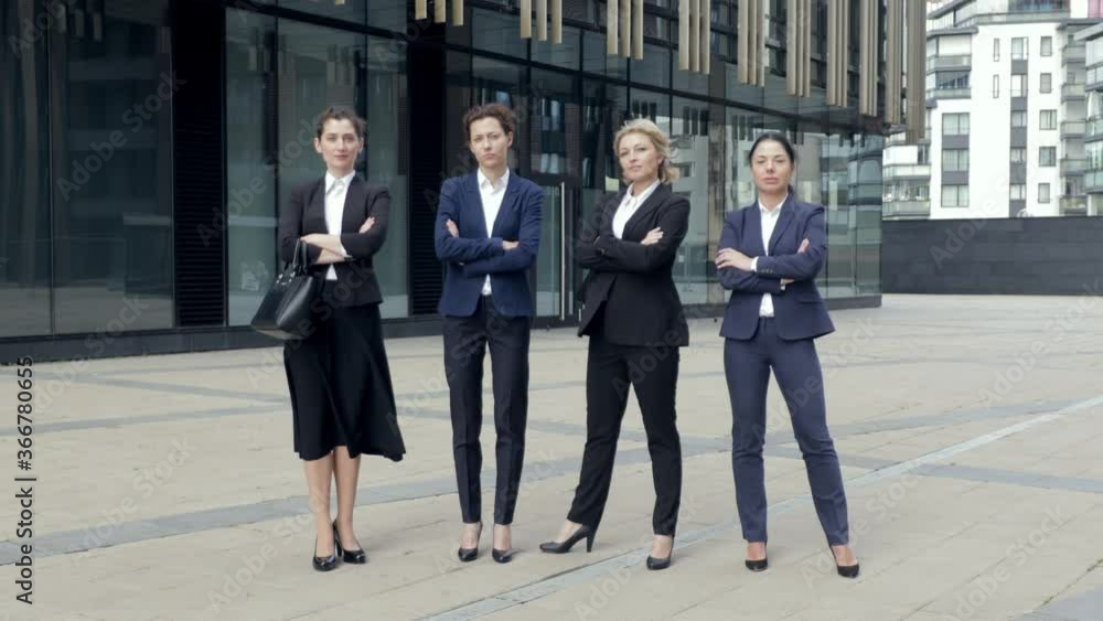 Serious company staff standing and crossing hands next to office. Group of successful women workers in suits looking to camera and smiling. Concept of team portrait. Serious company staff standing and crossing hands next to office. Group of successful women workers in suits looking to camera and smiling. Concept of team portrait.