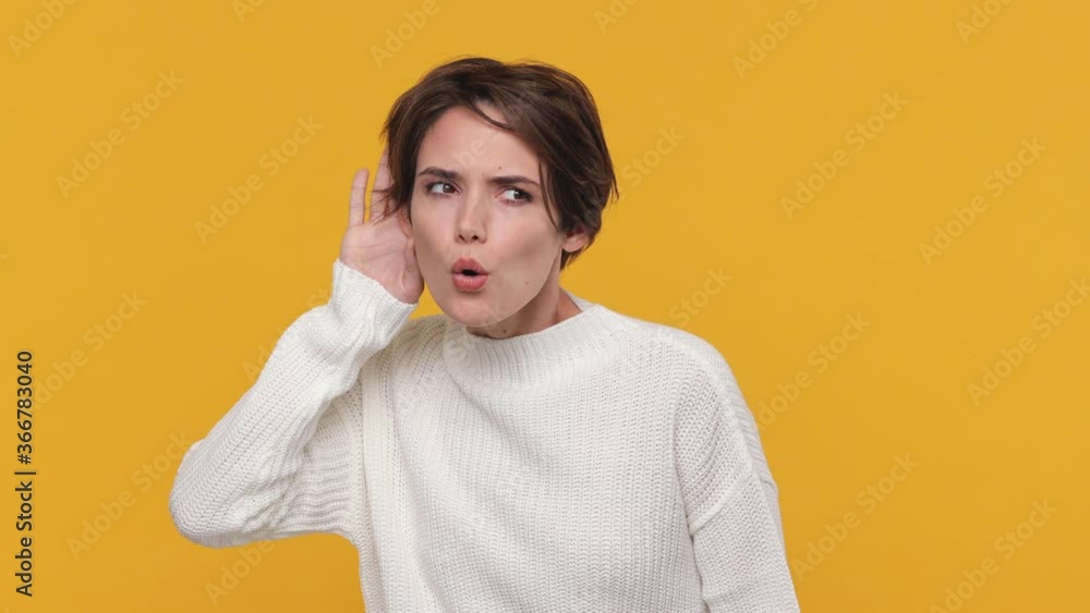 Fun beautiful young woman girl 20s years old in white sweater posing try to hear you overhear listening intently with hand near ear isolated on yellow background in studio. People lifestyle concept Fun beautiful young woman girl 20s years old in white sweater posing try to hear you overhear listening intently with hand near ear isolated on yellow background in studio. People lifestyle concept