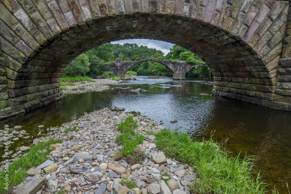 Fototapeta premium Cromwells bridge in the Ribble Valley, Lancashire. Old stone bridge over the river Hodder shot through an arch of the newer lower Hodder bridge.