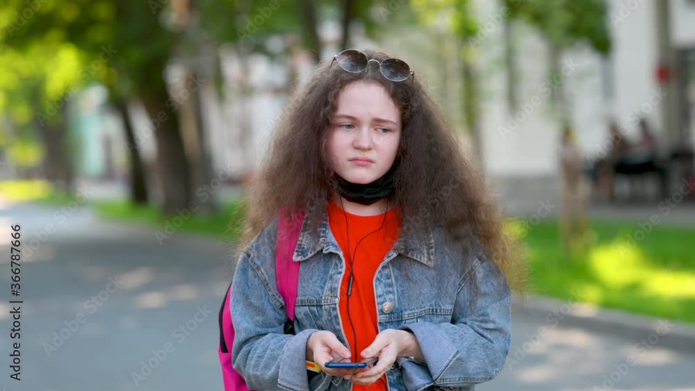 Young Attractive Girl in a Protective Mask on a City Street Communicates on a Smartphone. Takes off the Mask and is Sad. Young generation z interacts with phone in city uses app Young Attractive Girl in a Protective Mask on a City Street Communicates on a Smartphone. Takes off the Mask and is Sad. Young generation z interacts with phone in city uses app