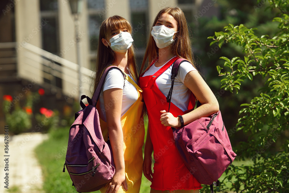 two teenage girls twins in a yellow and red school dress with purple backpacks outside the school in the afternoon on the lawn after the quarantine in masks