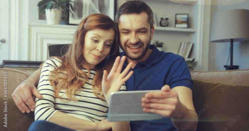 Happy married couple talking on video call from home using tablet. Two people laugh and chat with friends online in virtual app using webcam. People wave goodbye. Happy married couple talking on video call from home using tablet. Two people laugh and chat with friends online in virtual app using webcam. People wave goodbye.