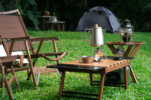 Stainless steel kettle, portable gas stove, bowl and vintage lanterns with outdoors table set on green lawn in camping area photo
