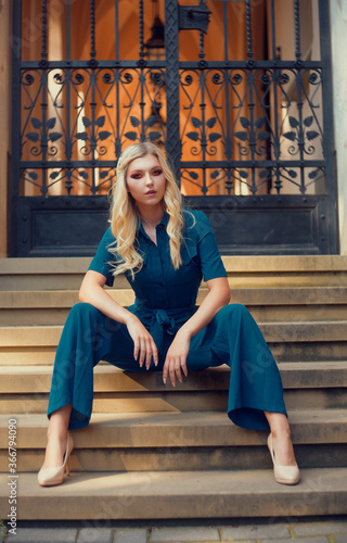 A full-length girl sits on the stairs in a long blue overalls. The girl looks into the camera with a daring look. Blonde with long legs poses.