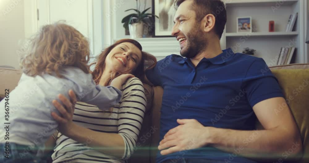 A little boy jumps on couch to his parents and indulges. Young parents are laughing with their youngest son. A little boy jumps on couch to his parents and indulges. Young parents are laughing with their youngest son.