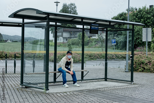 a young adult siting on a bench in a bus stop alone with a mask on waiting for the bus © Stavdahl