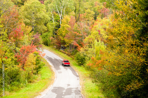 A winding road through a forest of hardwood trees showing autumn fall colors in Adirondack National Park in Upper New York. Red car is blurred to show motion.