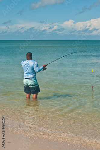 Back view, far distance of a African American, male, preparing to cast a fishing rod, with a fishing jig, while wading in calm, tropical water of the gulf of Mexico, on a sunny day