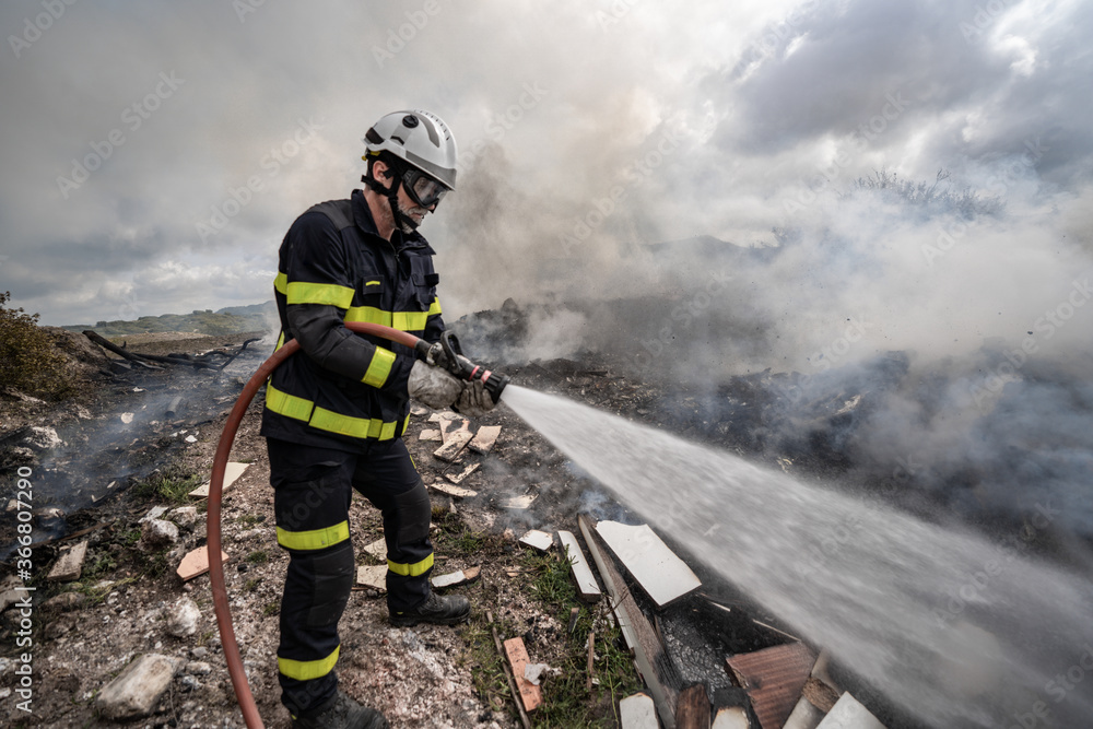 Side view of brave fireman in protective uniform standing with hose and extinguishing fire on dump in mountains Side view of brave fireman in protective uniform standing with hose and extinguishing fire on dump in mountains