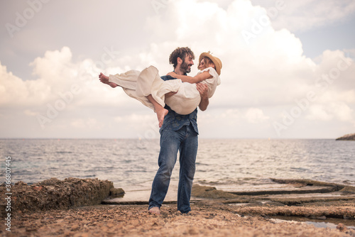 Low angle of smiling boyfriend lifting cheerful girlfriend above ground while standing on rocky coast near sea and looking at each other photo