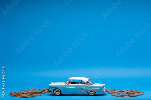 Light blue car figurine surrounded by a bunch of coins, on blue background.