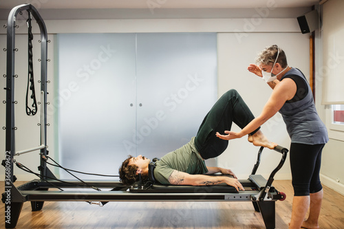 Concentrated male in sportswear doing exercises on pilates reformer during training under supervision of female instructor in protective respirator during coronavirus pandemic photo