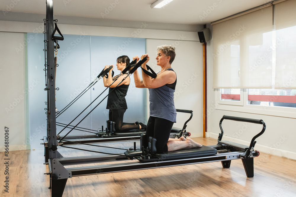 Side view of focused sportswomen in activewear doing exercises on pilates machine and pumping muscles with metal resistance equipment Side view of focused sportswomen in activewear doing exercises on pilates machine and pumping muscles with metal resistance equipment