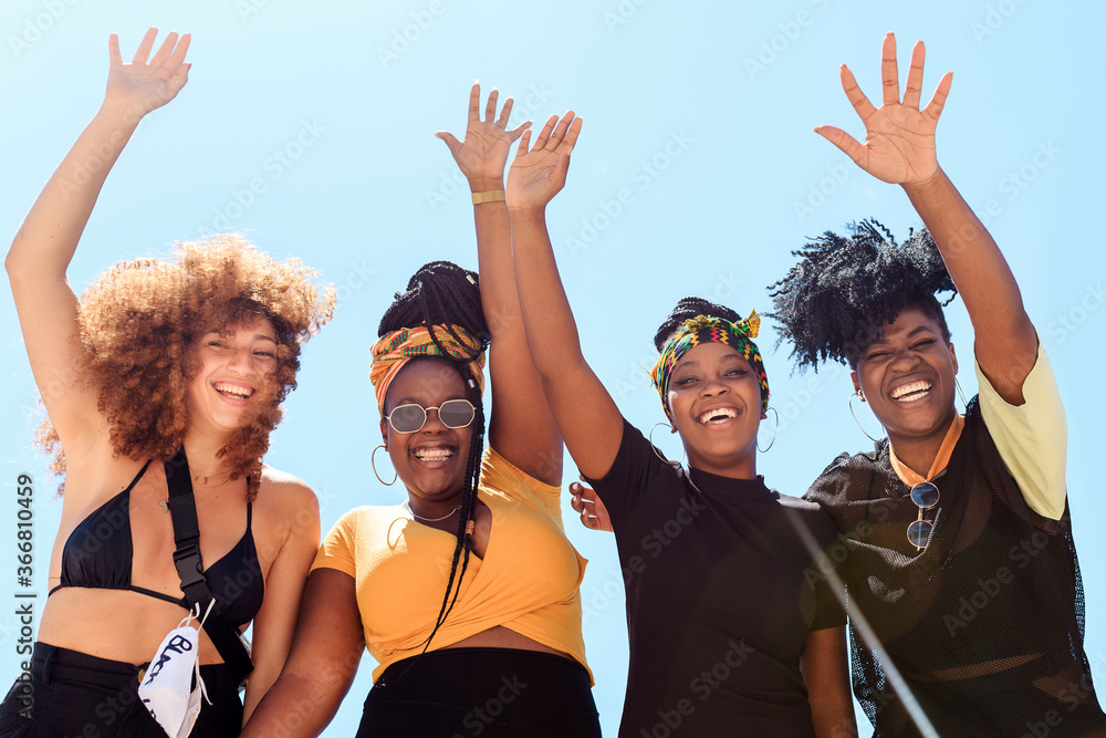 Low angle of company of friendly multiethnic females expressing joy with raised arms on background of cloudless sky in summer and looking at camera Low angle of company of friendly multiethnic females expressing joy with raised arms on background of cloudless sky in summer and looking at camera