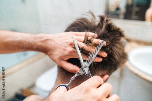 Back view of unrecognizable male making haircut to guy using scissors against blurred interior of light bathroom at home