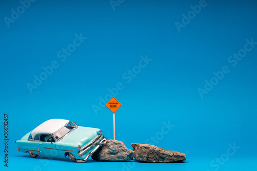 Light blue, retro car figurine next to two boulders and a construction zone sign, on blue background.