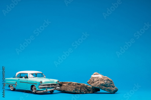 Light, blue retro car figurine next to two boulders, on blue background.