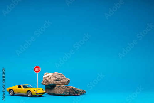 Yellow, sport car figurine next to two boulders and a stop sign, on blue background.