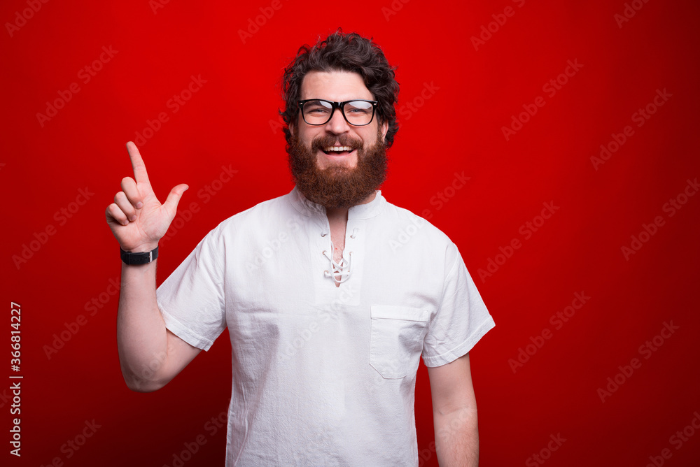 Smiling bearded man is pointing upwards while standing over red background. Smiling bearded man is pointing upwards while standing over red background.