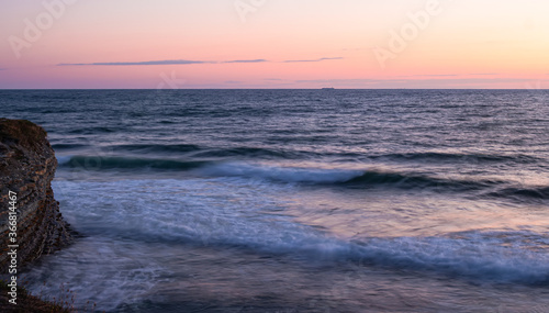 Soft pink sea sunset. Photo at a long shutter speed of one second. Smooth waves beat against the rocks. Cargo ships and tiny clouds can be seen on the horizon.